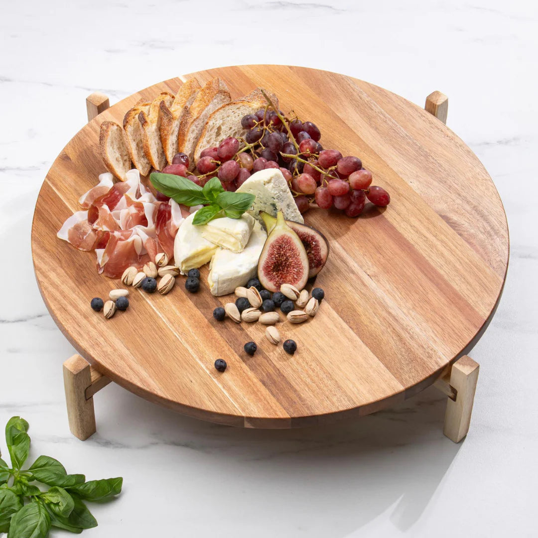 Gather Elevated Lazy Susan. Wooden round cheese board with assorted fruits, meats, and cheeses on a white marble surface.