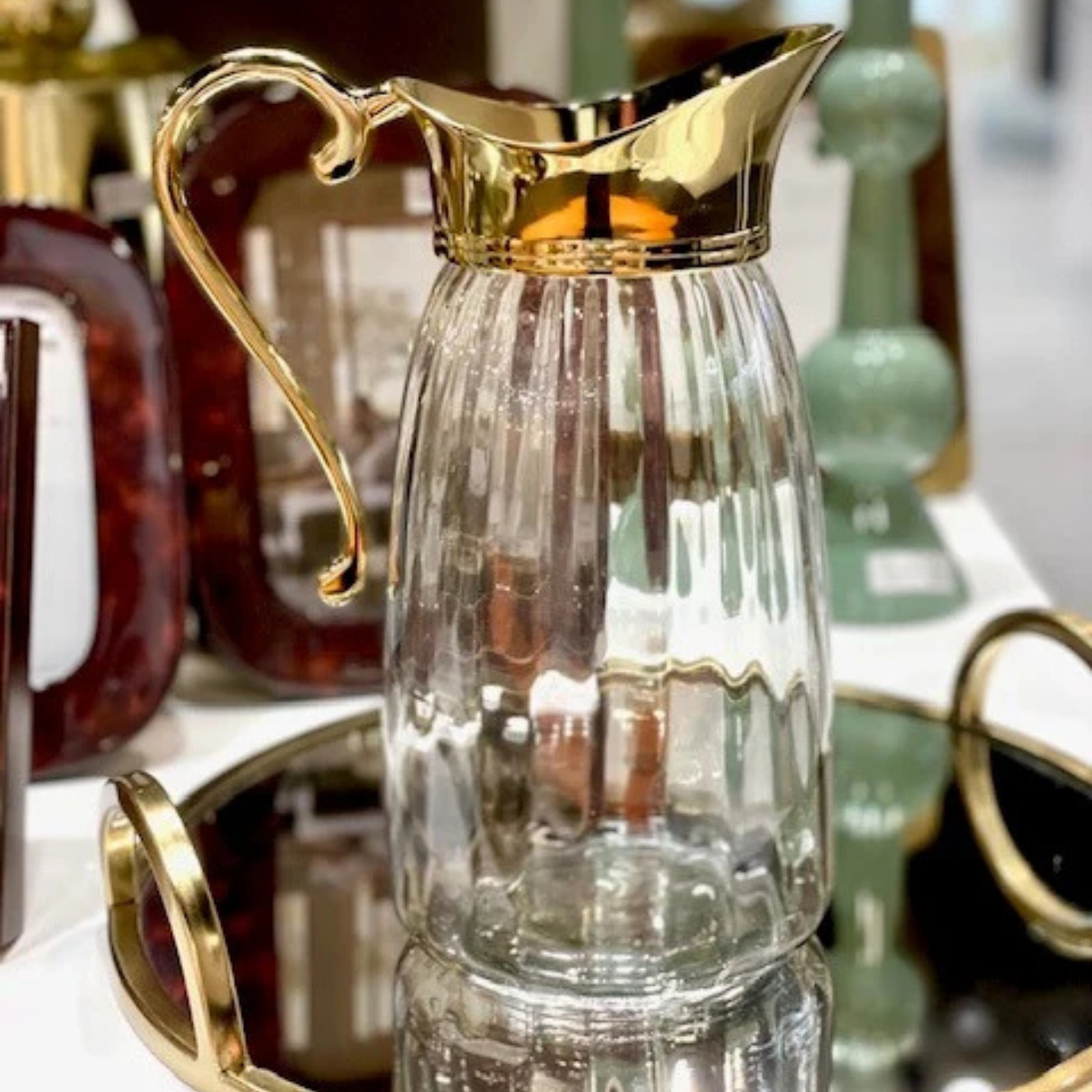A clear ribbed glass pitcher with a shiny gold metal handle and pouring spout, displayed on a reflective tray with other decorative items in the background.