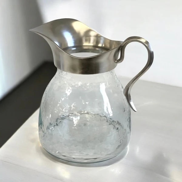 Clear glass pitcher with metal lid and handle on a white surface