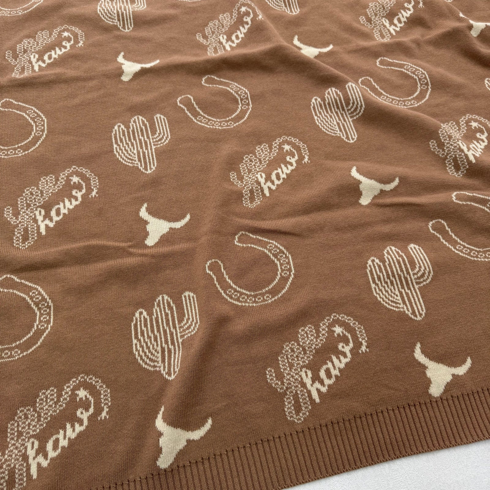 A close-up of a brown knit fabric with a repeating white pattern of cacti, horseshoes, longhorn silhouettes, and the word 'hows'.
