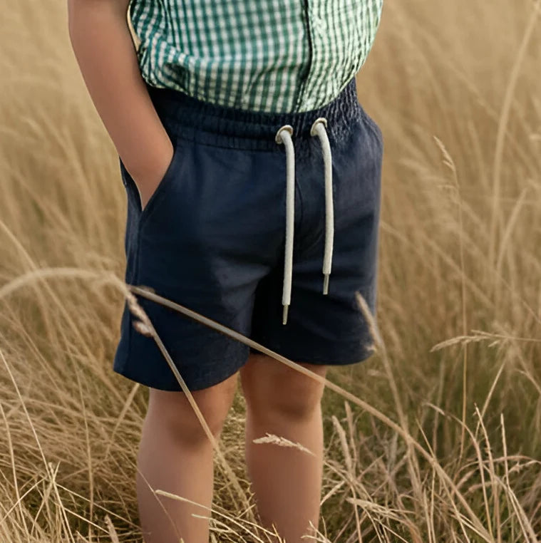 A child wearing navy blue shorts with a white drawstring and a green-and-white gingham shirt, standing in a field of dry golden grass.