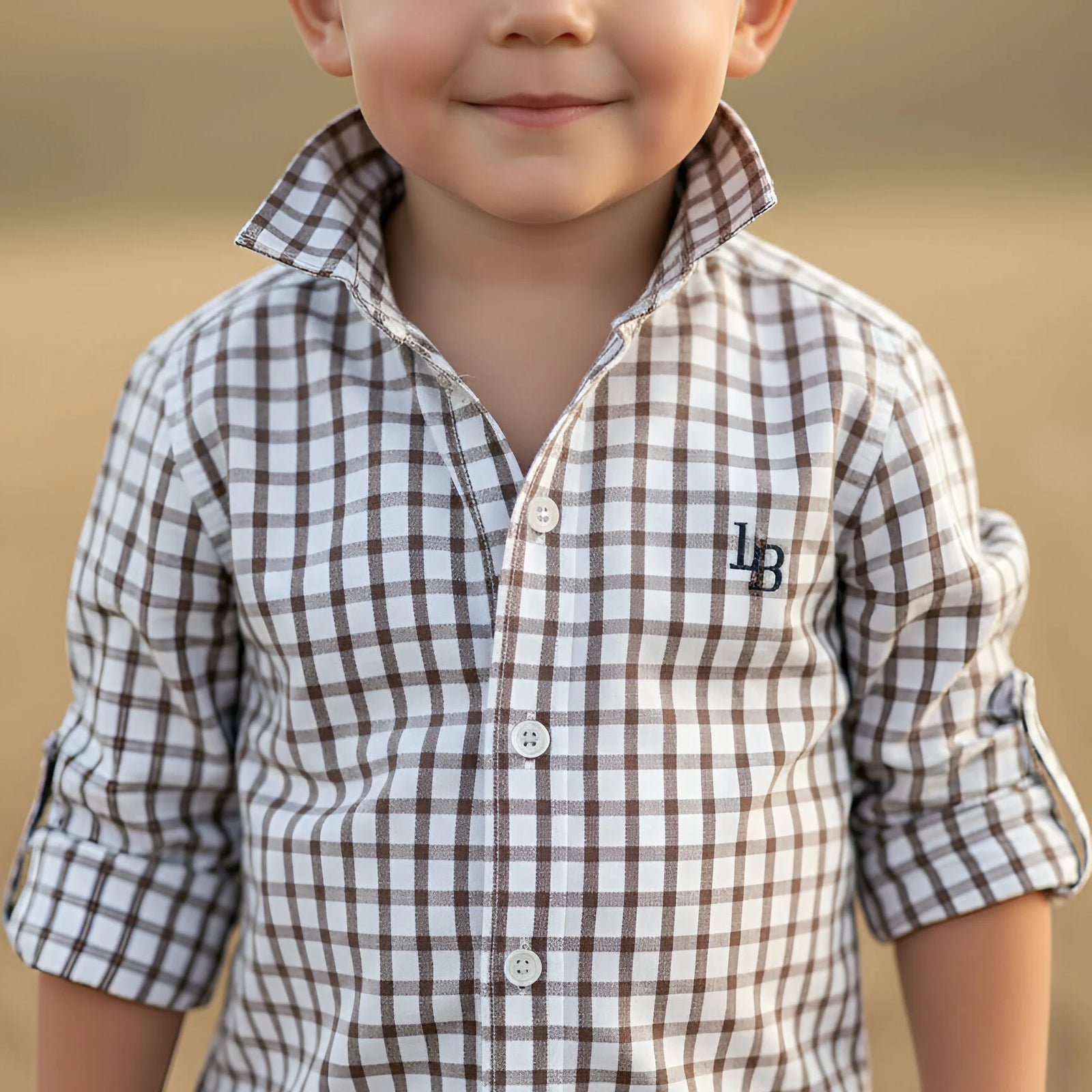 A close-up of a young boy wearing a brown and white checkered button-down shirt with a collar and rolled-up sleeves. The shirt features an embroidered monogram 'LB' on the left chest.