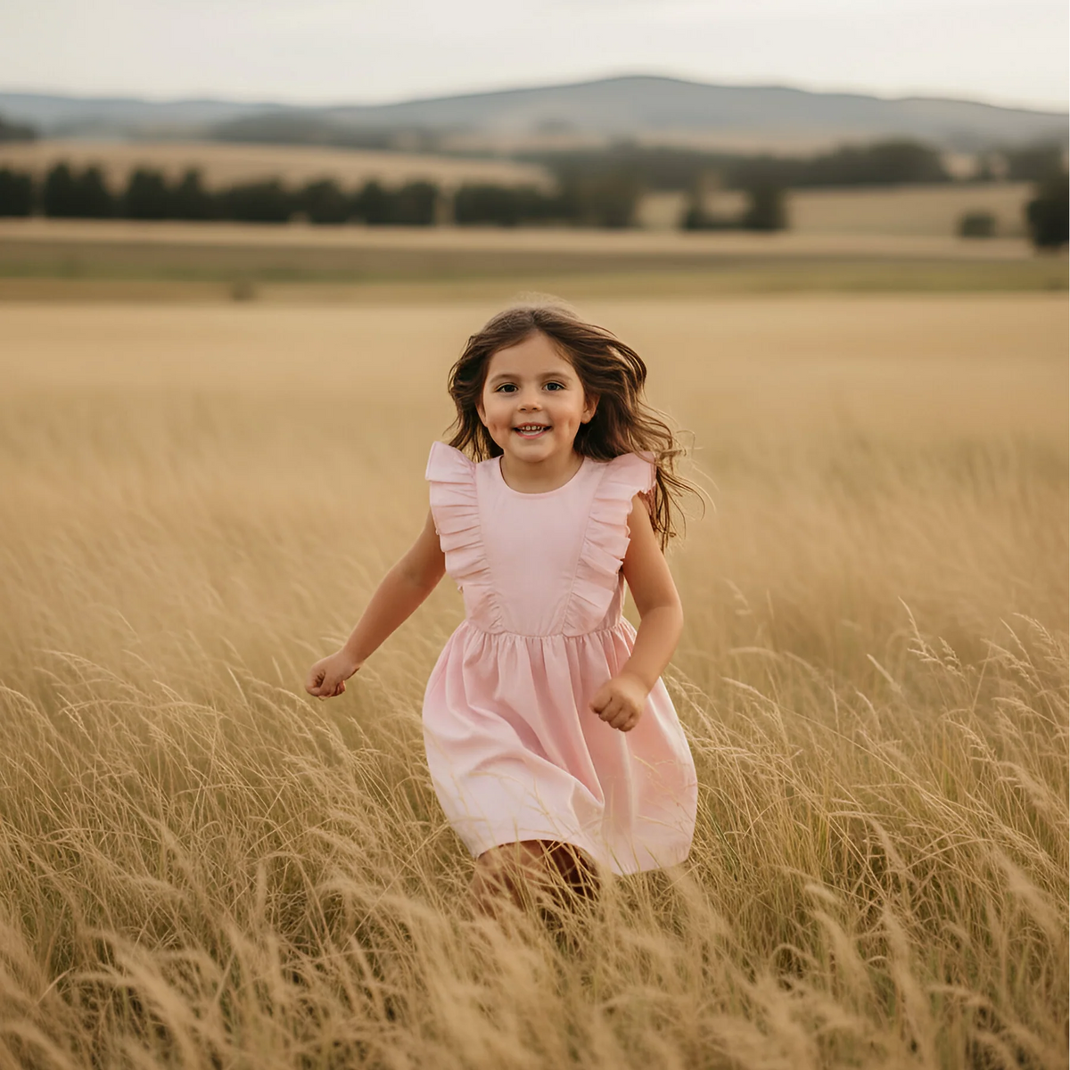 A young girl with long brown hair runs through a golden wheat field wearing a light pink ruffle-sleeve dress. The background features a soft-focus landscape of rolling hills under a warm, golden sky.