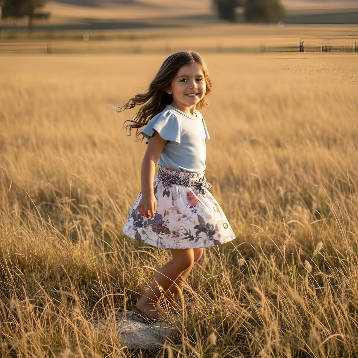A young girl with long brown hair stands in a golden grassy field at sunset, wearing a light blue short-sleeve top and a white floral skirt with a patterned waistband.