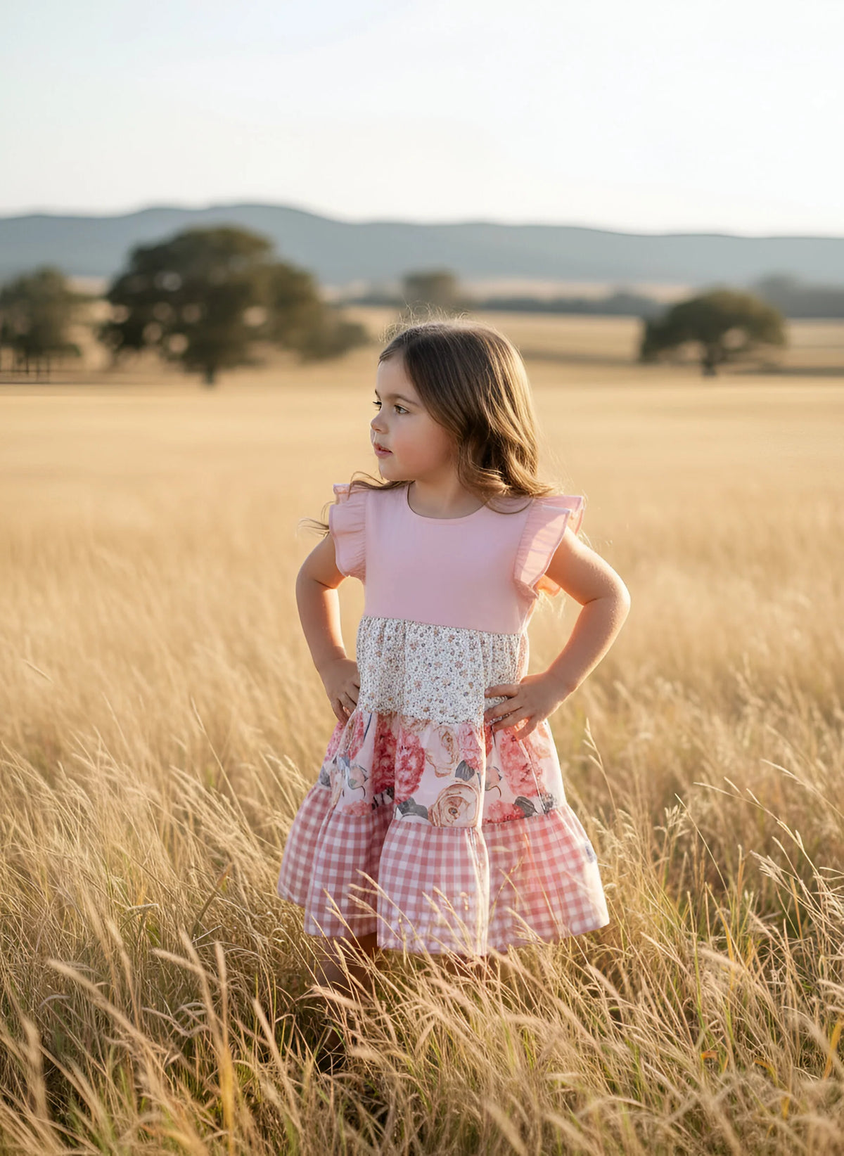 A young girl stands in a golden wheat field at sunset wearing a tiered dress with a pink bodice, floral lace panel, and pink-and-white gingham skirt.