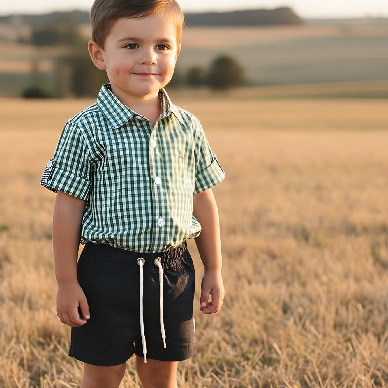 A young boy standing in a sunlit field, wearing a green and white gingham short-sleeve button-up shirt and dark navy shorts with white drawstrings.