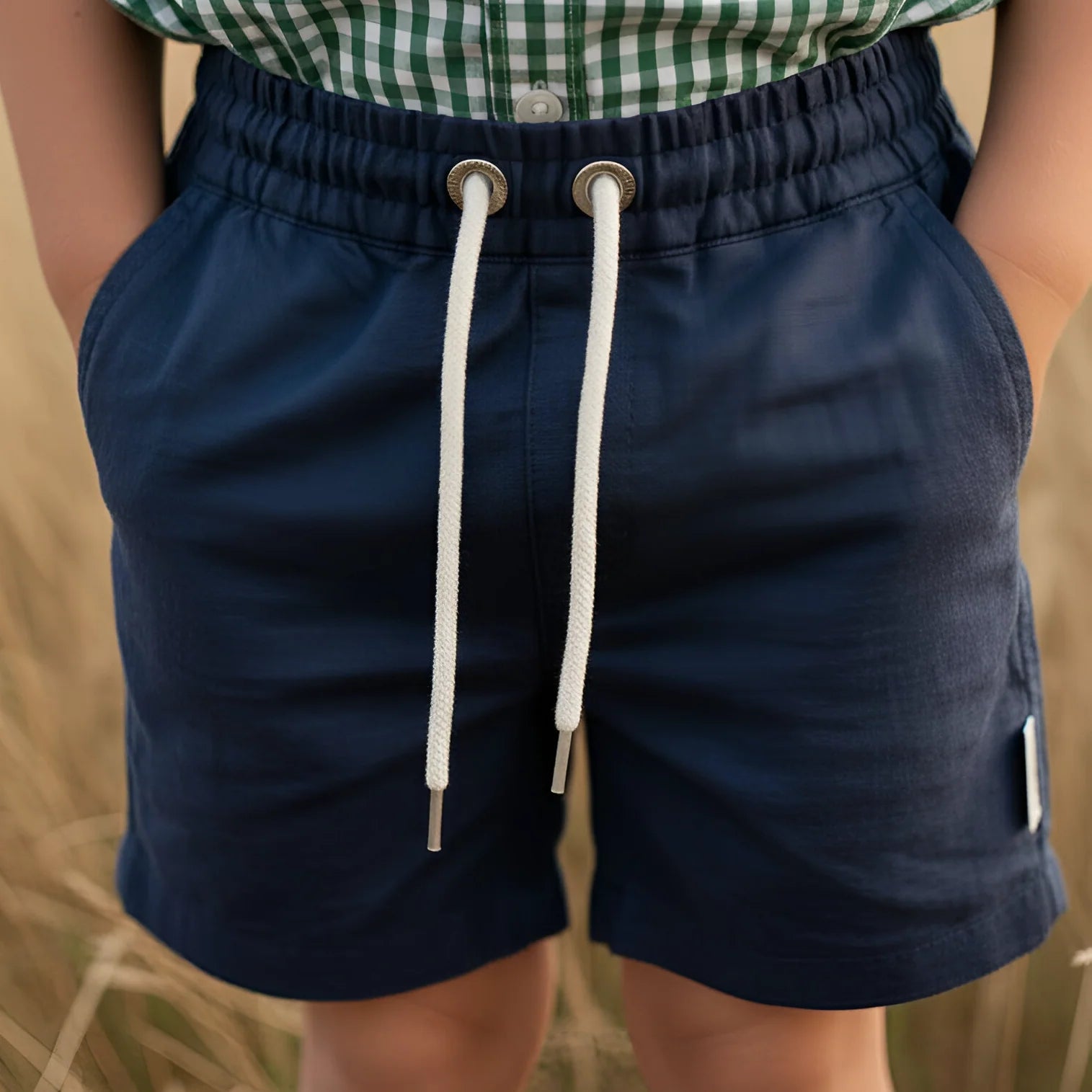 A young boy standing in a sunlit field, wearing a green and white gingham short-sleeve button-up shirt and dark navy shorts with white drawstrings.