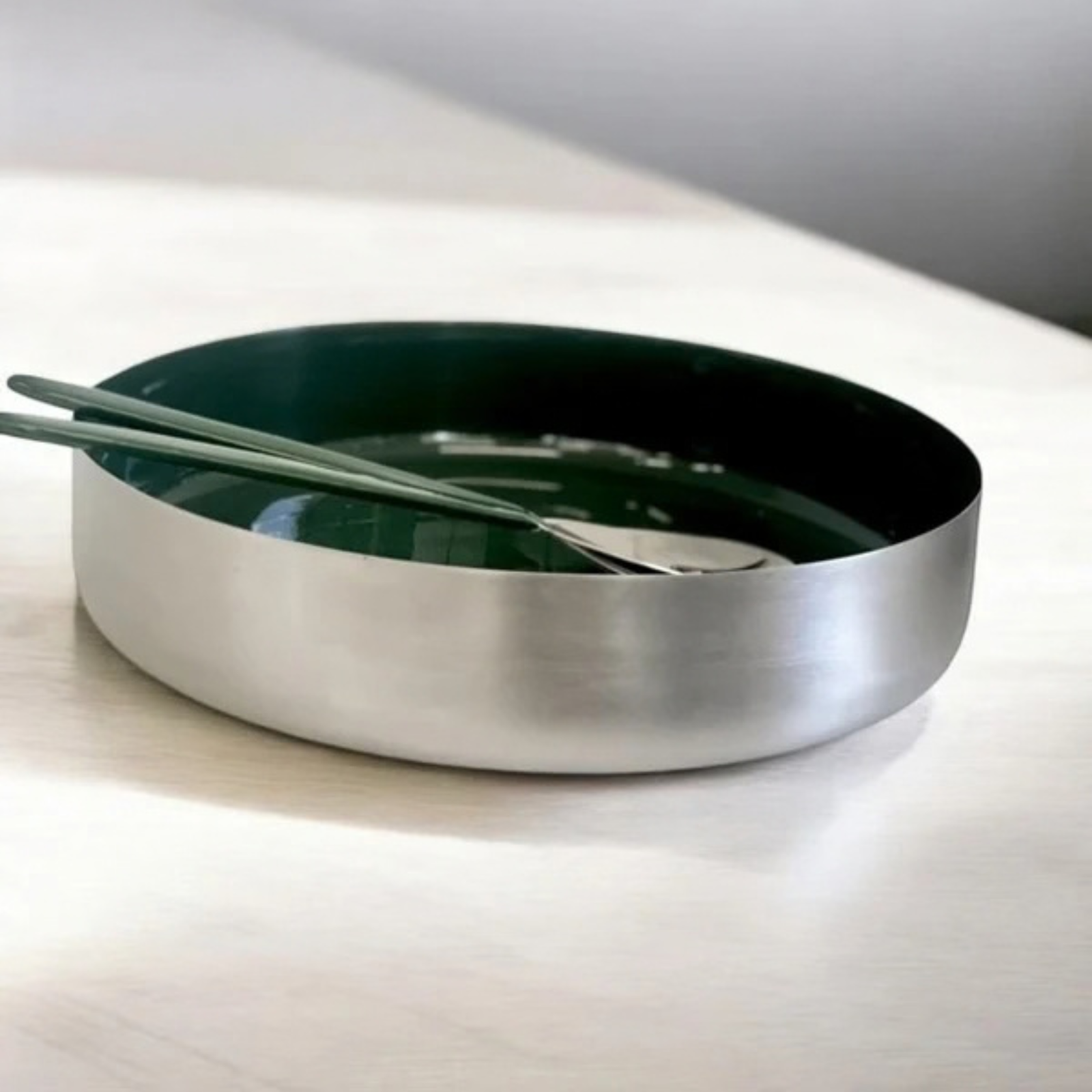 A modern round metal bowl with a brushed silver exterior and a dark green interior, shown on a light wood surface with chopsticks and a spoon resting inside.