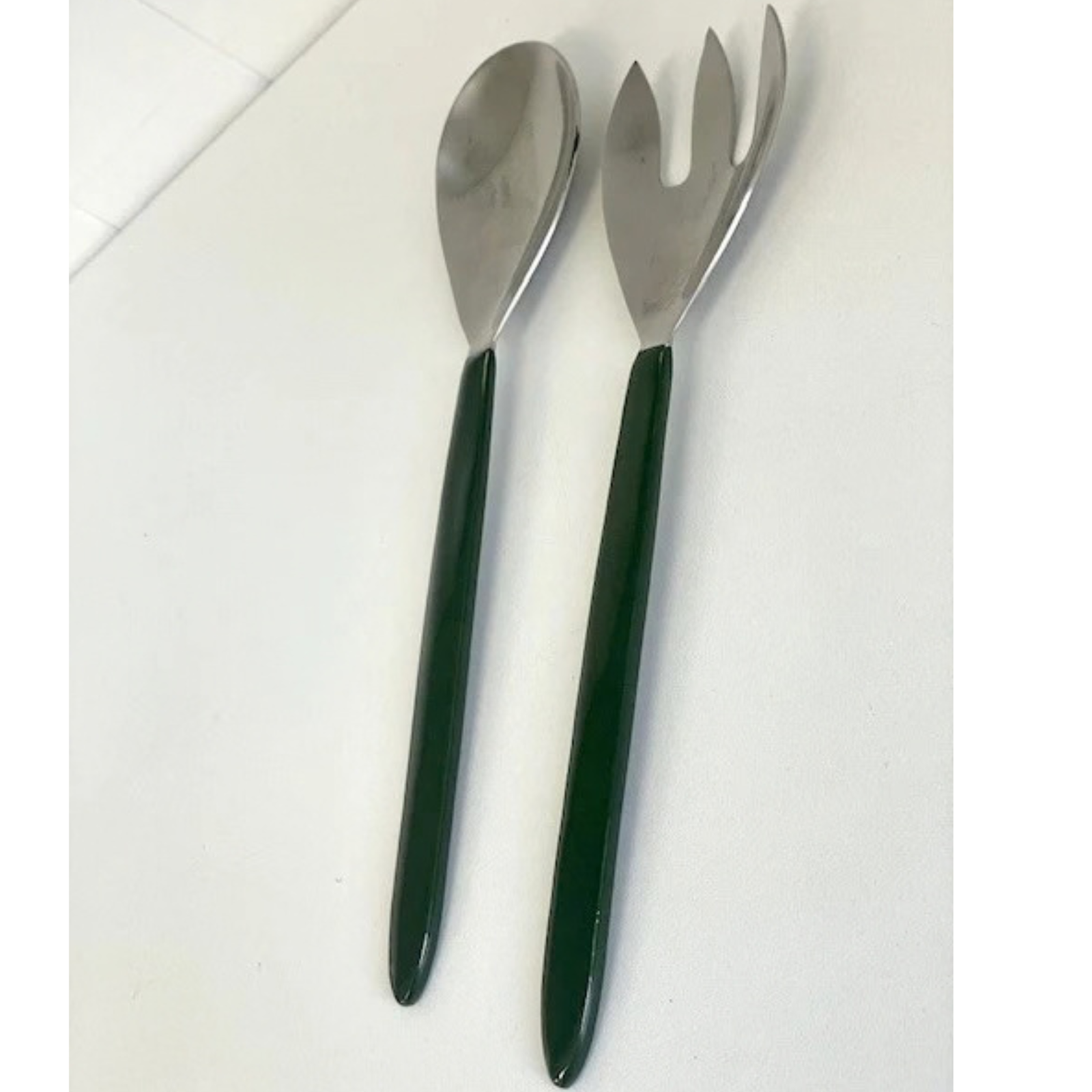 A stainless steel salad server and serving fork set with dark green handles, displayed on a light off-white surface.