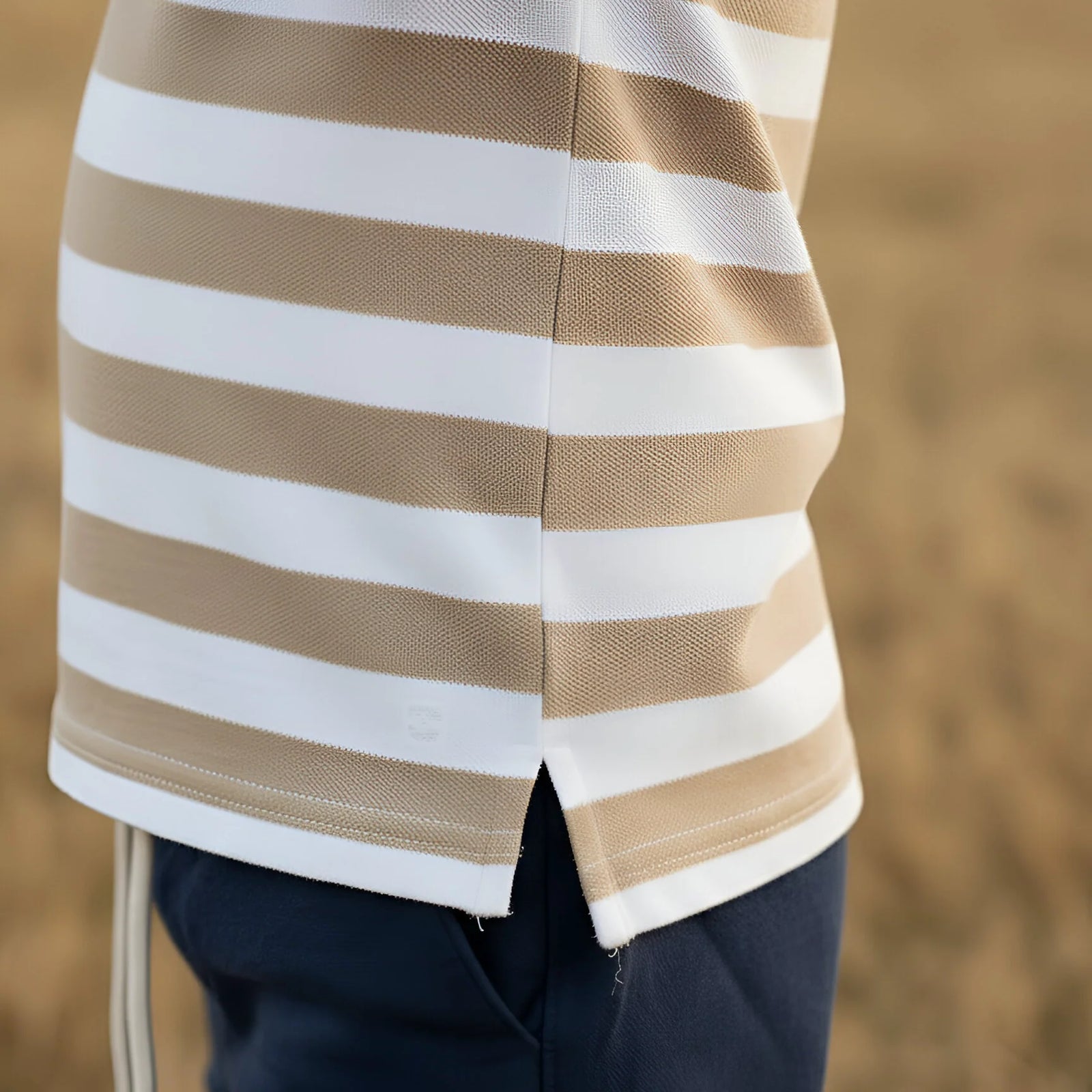 Close-up of a person wearing a beige and white horizontally striped knit top with a split hem, paired with dark navy pants, against a blurred outdoor background.