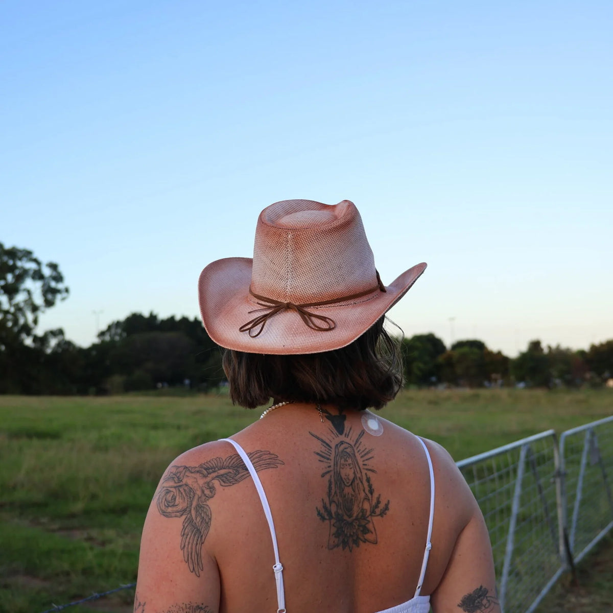 Person wearing a cowboy hat with tattoos on their back, standing in an open field.