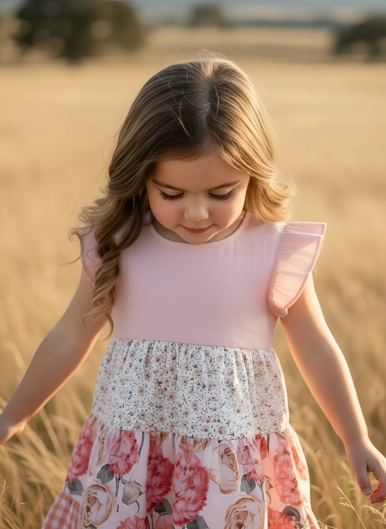A young girl with long light brown hair stands in a golden field wearing a pink sleeveless dress with ruffled shoulders, a white lace waistband, and a pink floral skirt.