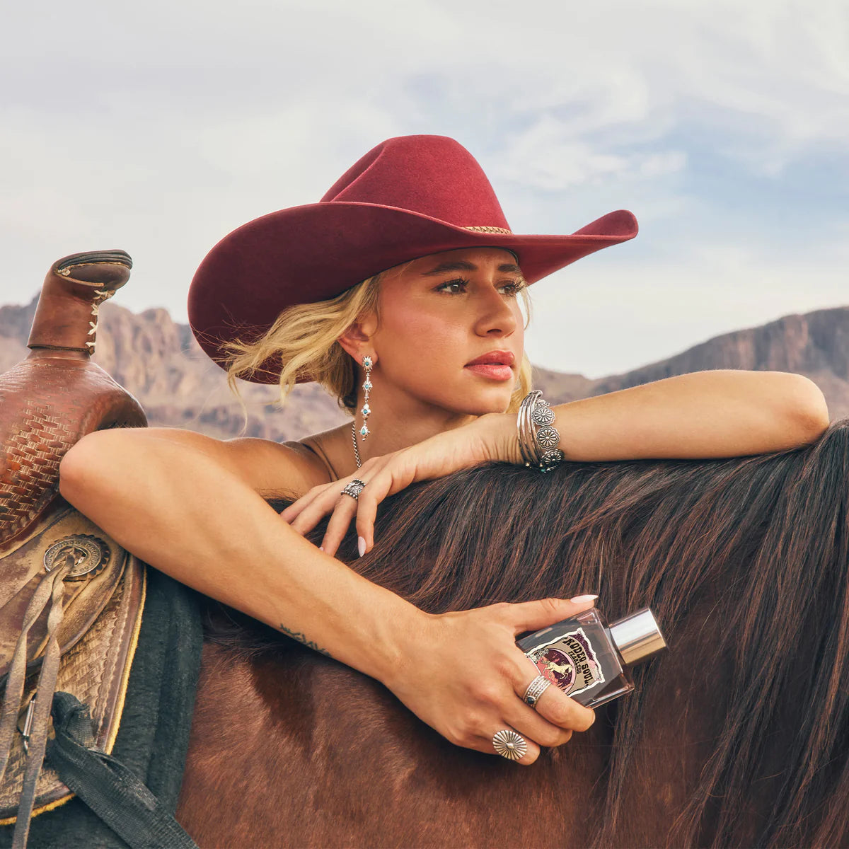 Tru Western Rodeo Soul Fearless Eau De Parfu. Woman in a cowboy hat holding a bottle of Jack Daniel's whiskey on a horse with mountains in the background