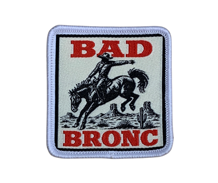 A square embroidered patch featuring a black cowboy riding a bucking horse, with the words 'BAD BRONC' in red text on a light gray background, finished with a white stitched border.