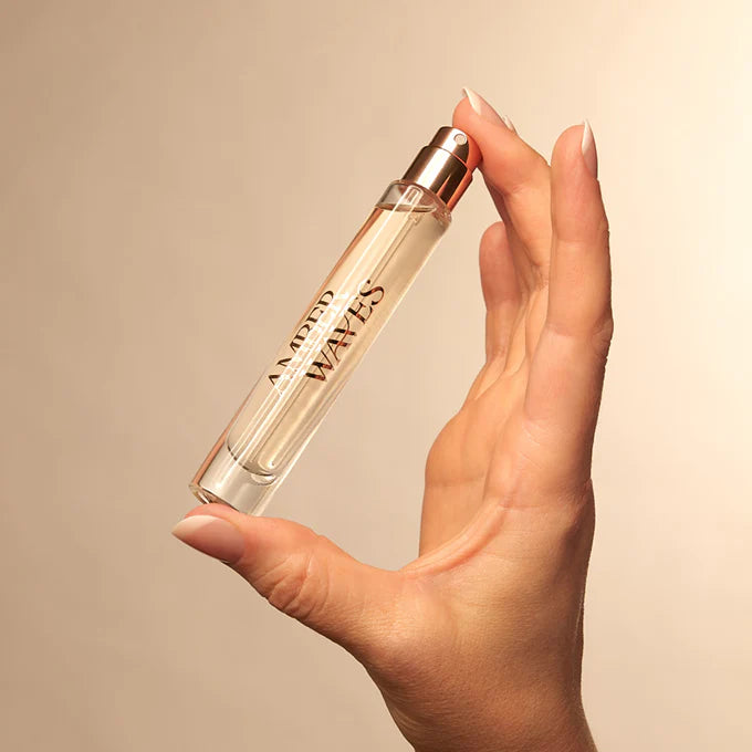 Hand holding a transparent perfume bottle with a gold cap against a beige background