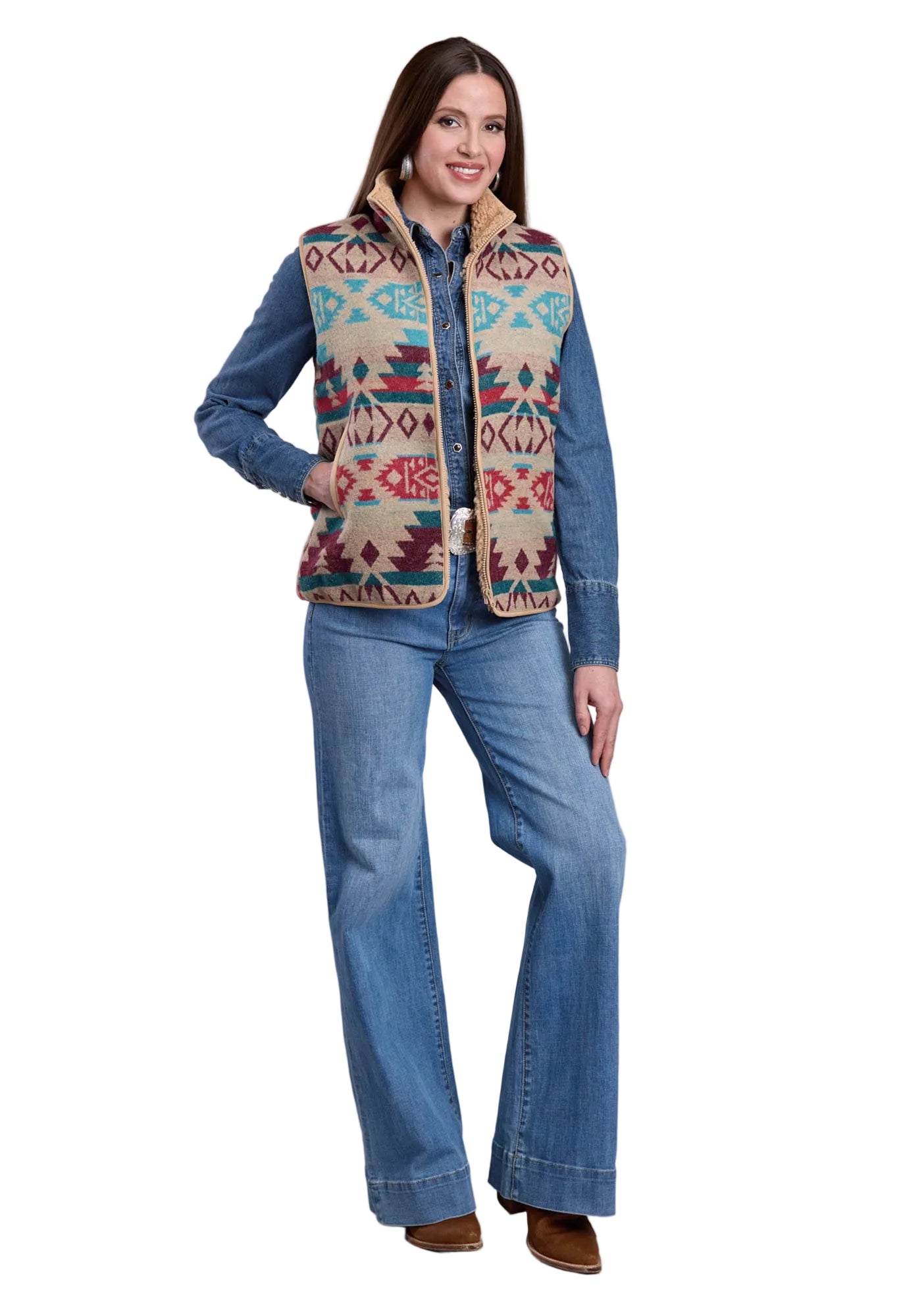 Woman wearing a patterned vest over a denim shirt with jeans on a plain background