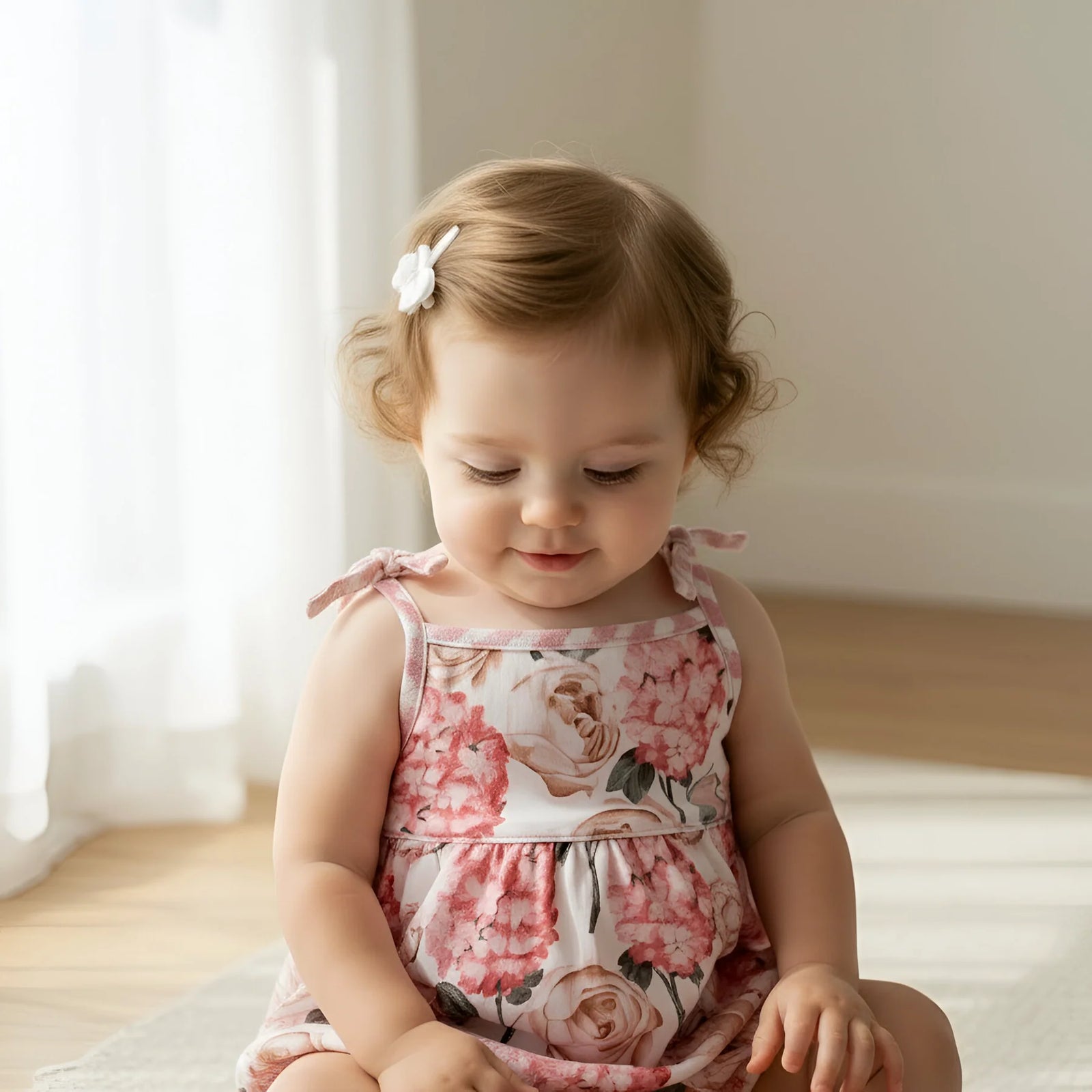 A toddler girl with light brown hair wearing a pink and white floral romper dress, sitting on a rug in a bright, softly lit room.
