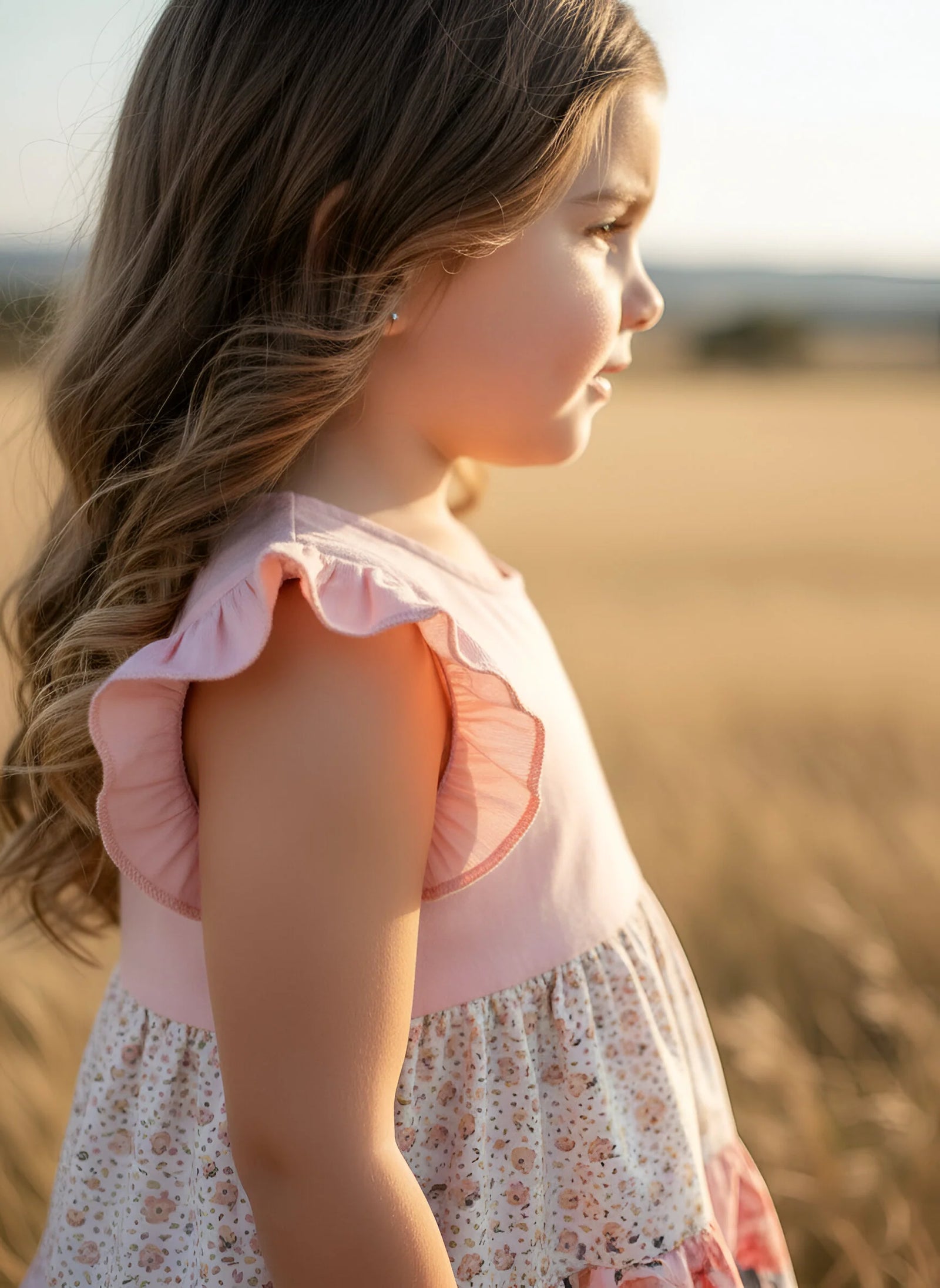 A young girl in profile wearing a light pink and white floral dress, standing in a warm, golden field.