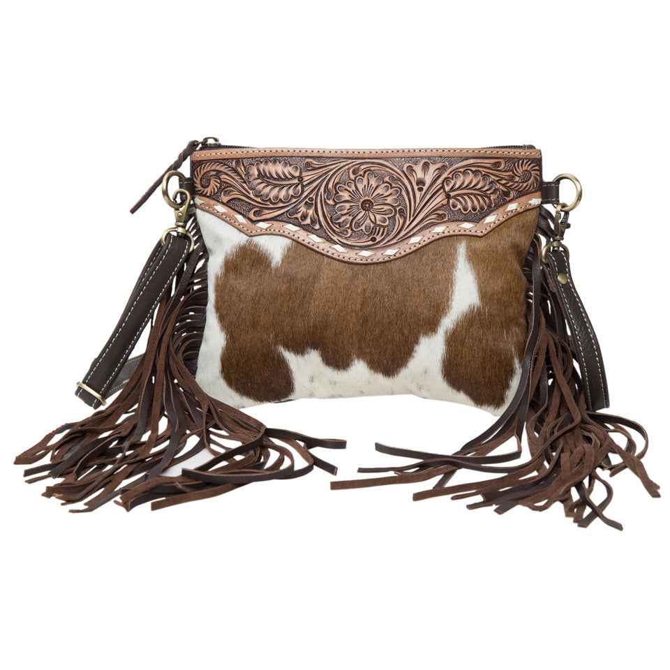 The Design Edge Women's Curico Cowhide Bag. An oversized cowhide clutch bag with hand tooled details and leather fringes, in brown and white colors, featuring a top zipper closure and side tassel details.