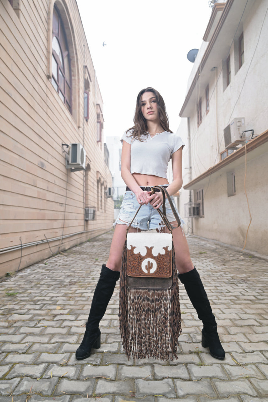 Women's Tooling Leather Flap Sling Cowhide Bag With Fringes. Woman holding a brown handbag with white floral design on a cobblestone street.