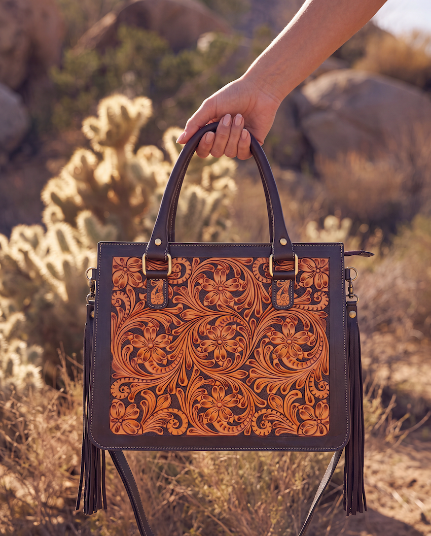 A hand holds a structured brown leather handbag with an intricate floral tooled pattern on the front, set against a desert landscape with cacti and rocks.