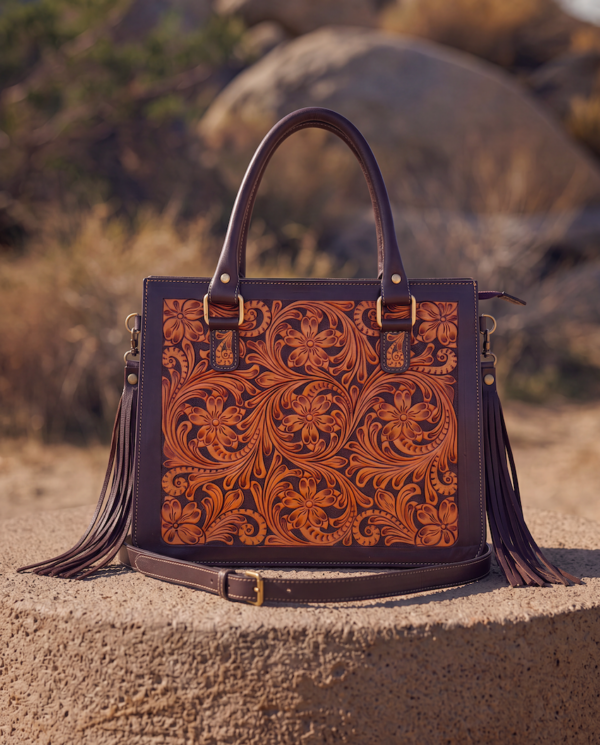 A structured brown leather handbag with an intricate tooled floral pattern on the front, dual top handles, and long leather tassels on the sides, displayed on a stone pedestal in an outdoor desert setting.