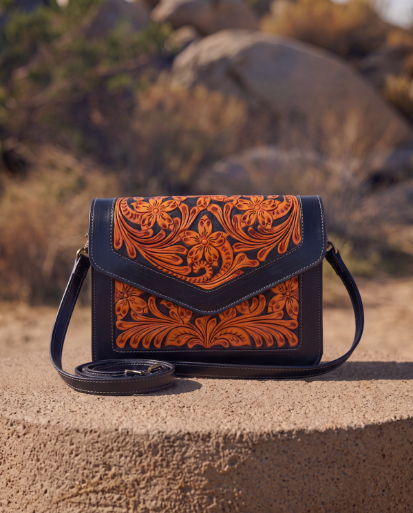 A hand-tooled leather crossbody bag with a dark navy blue base and vibrant orange floral carvings, resting on a stone surface with a blurred outdoor desert landscape in the background.