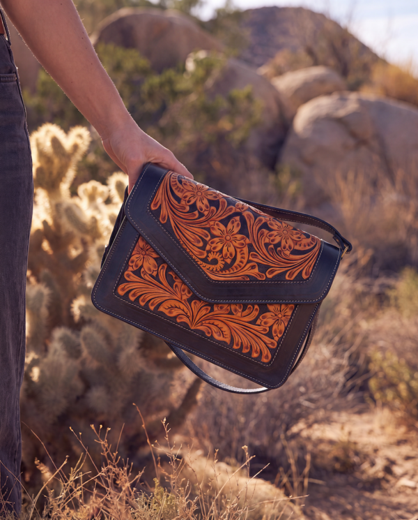 A hand holding a black leather shoulder bag with intricate orange tooled floral patterns, set against a desert landscape with cacti and rocks.
