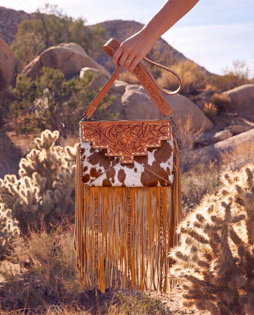 A hand holds a western-style fringe bag outdoors in a rocky desert landscape. The bag has a tan tooled leather top, a brown-and-white cowhide front panel, and long tan fringe hanging below.