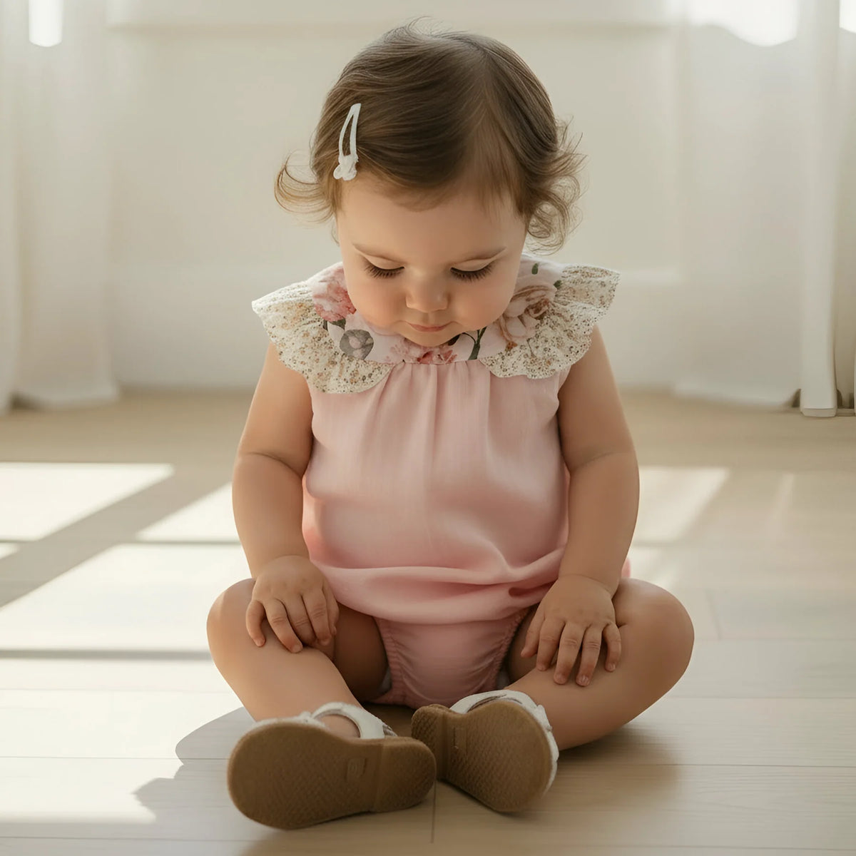 Baby sitting on a wooden floor wearing a pink outfit with lace details.