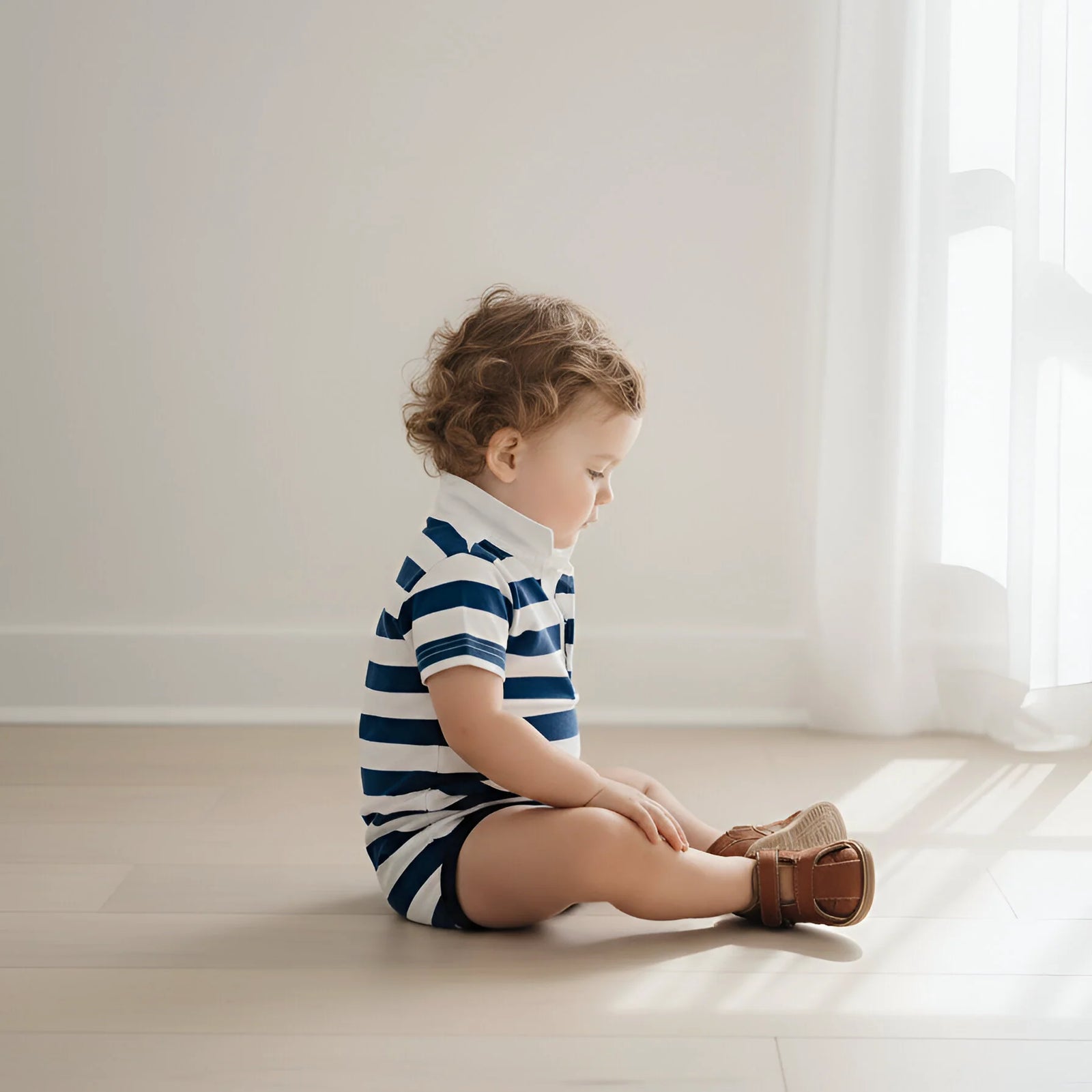 A toddler sitting on a light wooden floor in a bright room, wearing a navy and white horizontally striped short-sleeve romper and brown leather shoes.