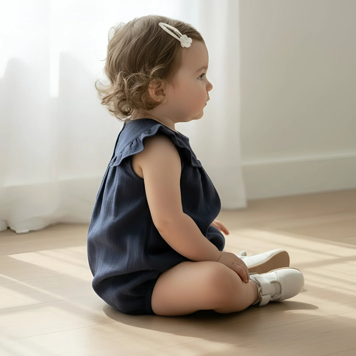 A toddler girl with curly light brown hair sits on a light wood floor wearing a sleeveless navy blue romper, white shoes, and a small white hair clip, looking to the side in a bright room.