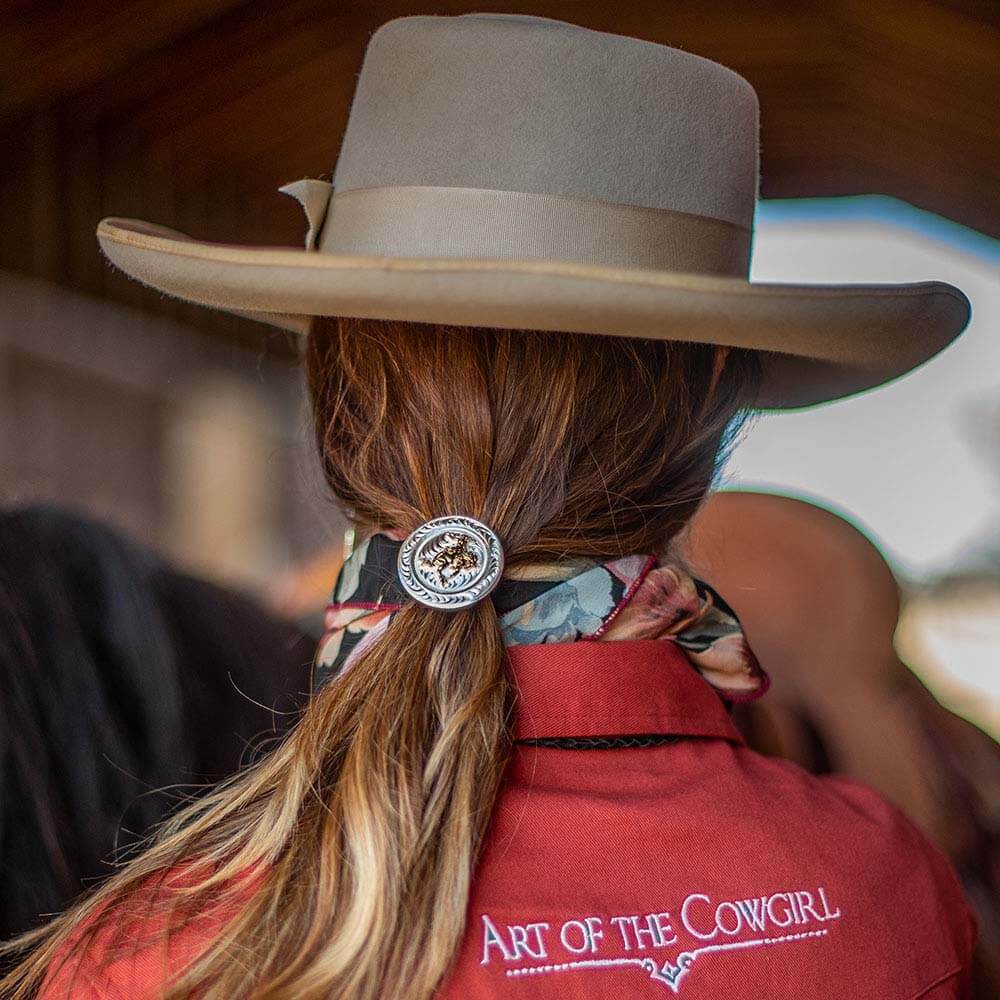 Montana Silversmiths Ponytail Holder. A round silver concho with an engraved design and an antiqued gold figure of a cowgirl on a bucking horse in the center, attached to a black ponytail holder with silver-toned tips.