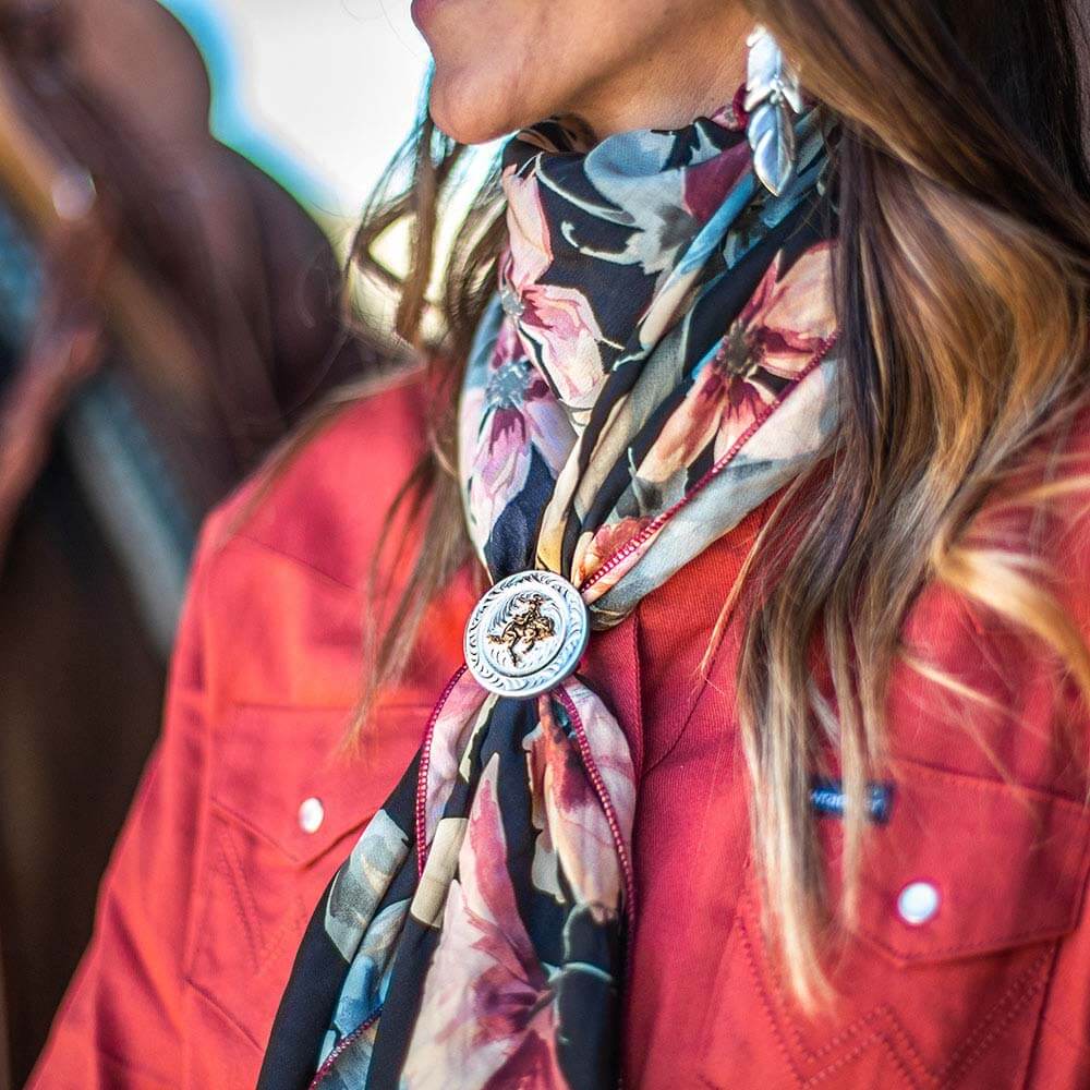 Montana Silversmiths Ponytail Holder. A round silver concho with an engraved design and an antiqued gold figure of a cowgirl on a bucking horse in the center, attached to a black ponytail holder with silver-toned tips.