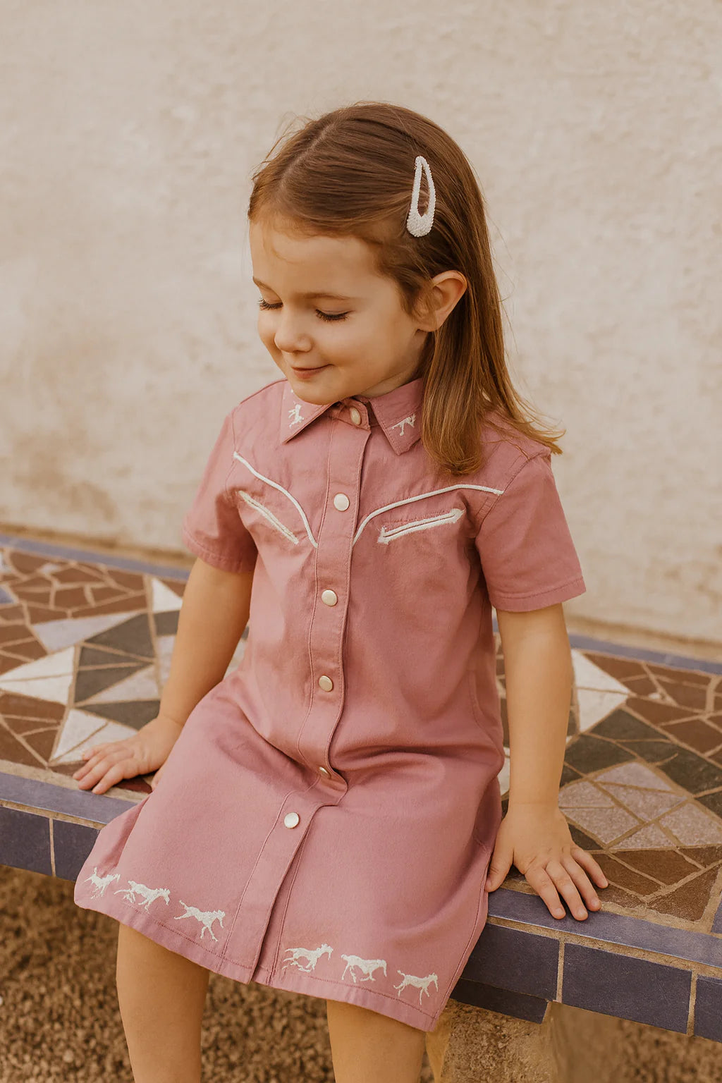 Little Windmill Girl's Arizona Western Blush Pink Dress. Young girl in a pink dress with horse embroidery sitting on a mosaic-tiled floor.