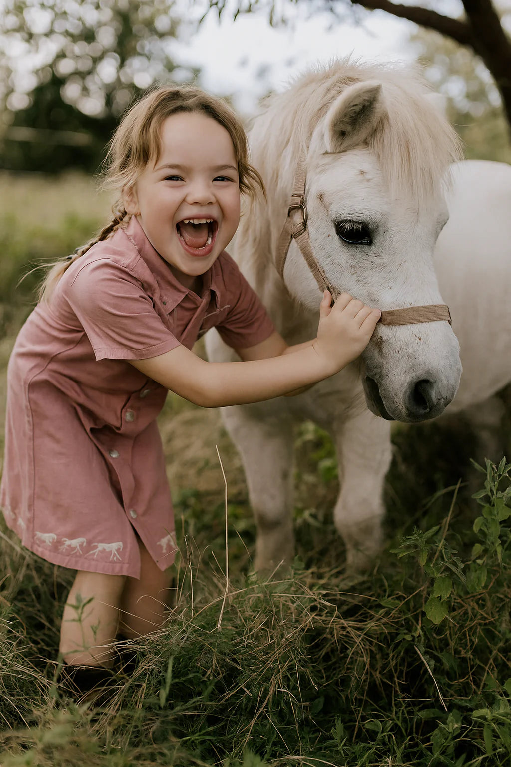 Little Windmill Girl's Arizona Western Blush Pink Dress. Young girl in a pink dress smiling and petting a small white horse in a grassy field.