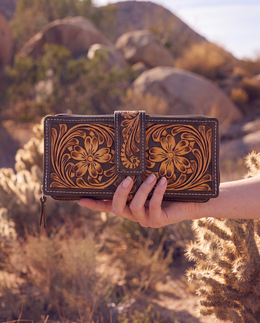 A hand holds a tooled leather wallet in a desert outdoor setting with rocks and cacti in the background. The wallet features intricate floral and scroll tooling in warm tan and dark brown tones.