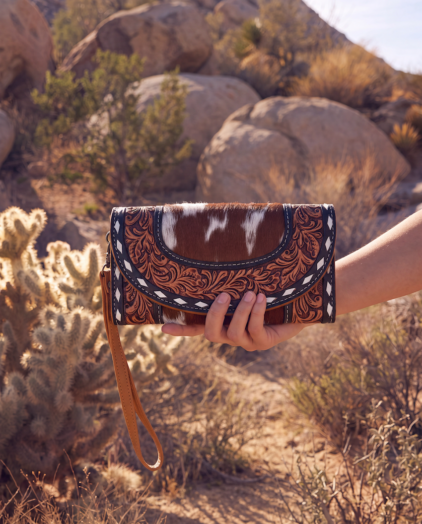 A hand holding a western-style clutch purse with a brown and white cowhide pattern, tooled leather detailing, and a wrist strap, set against a desert landscape with rocks and cacti.