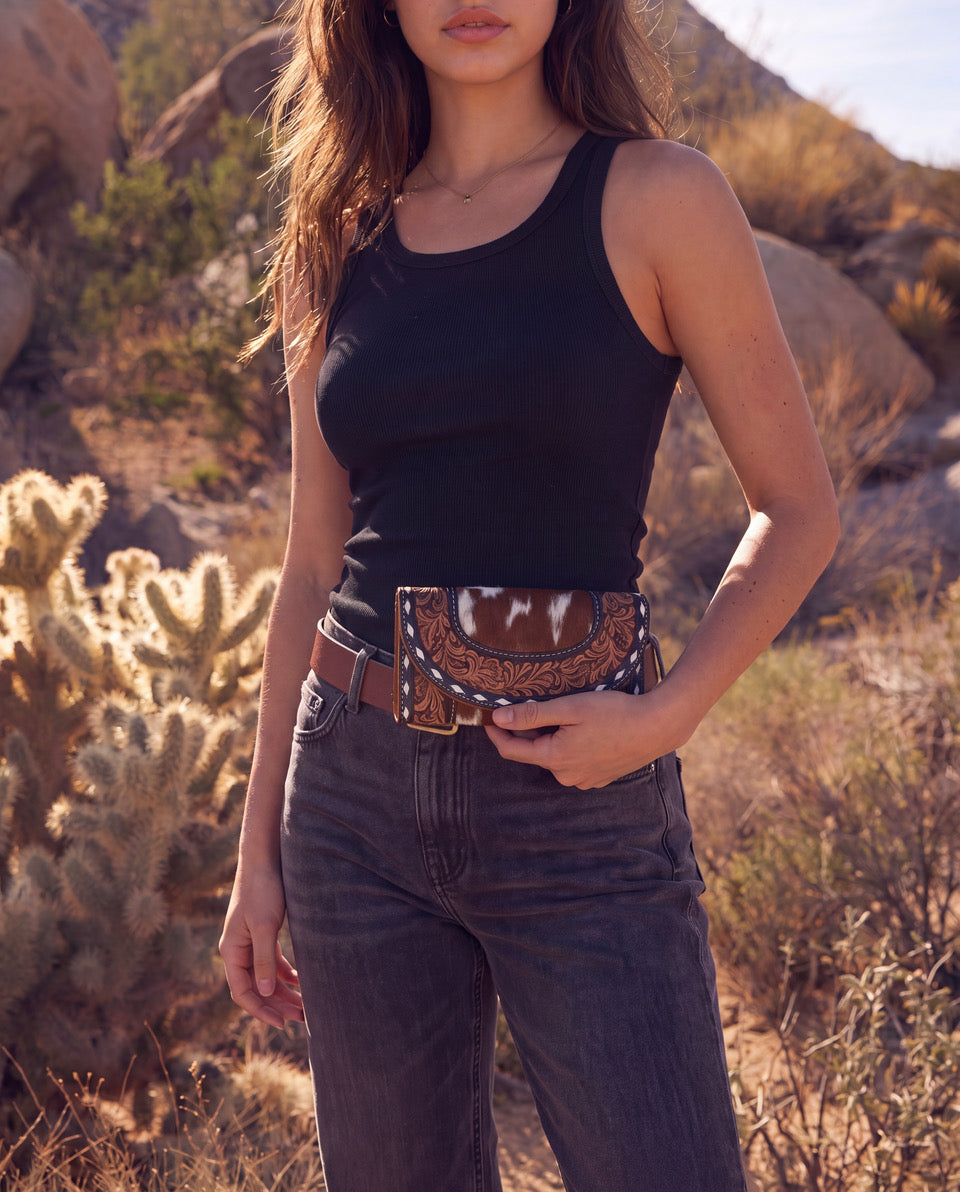 A woman wearing a black tank top, dark gray jeans, and a brown western belt with a decorative horn buckle, standing outdoors in a desert landscape with cacti and rocks.