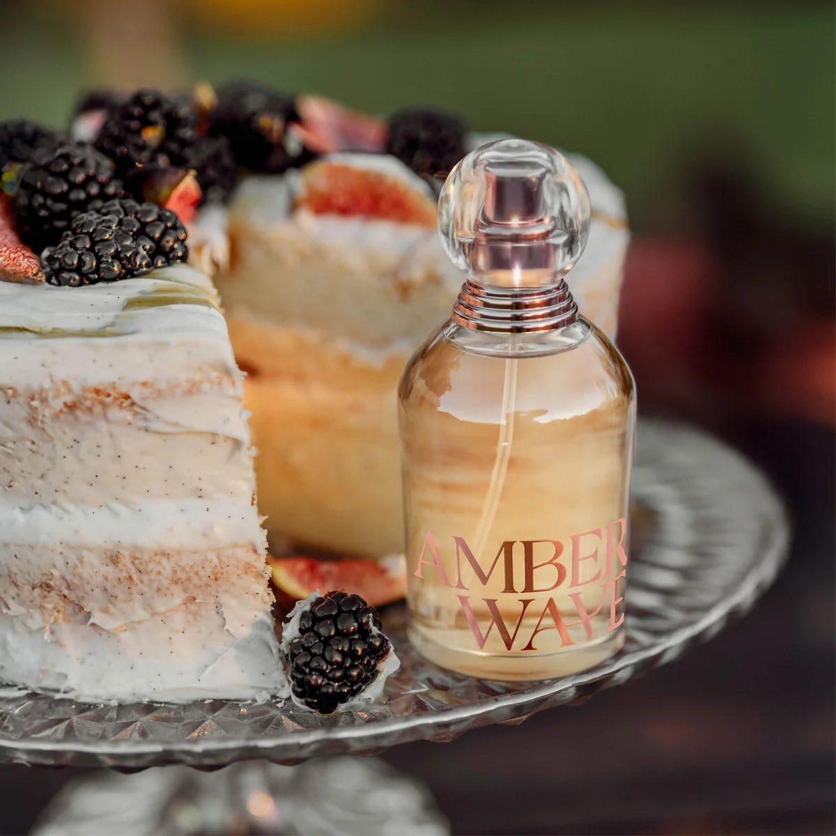 Perfume bottle labeled 'Amber Wave' on a cake stand with a cake and berries in the background