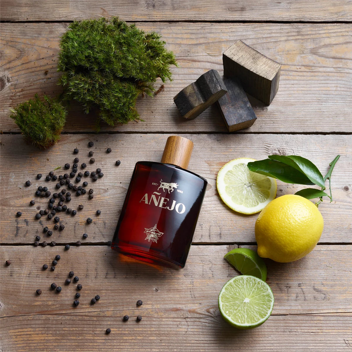 A bottle of Anejo aged rum spirit on a rustic wooden surface, surrounded by ingredients that define its flavor profile, including fresh lemons, whole black peppercorns, green moss, and dark wood blocks.