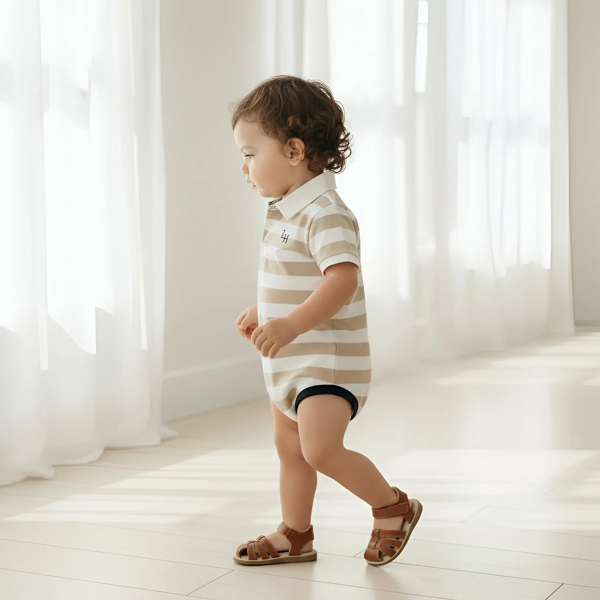 A toddler walking indoors wearing a short-sleeve polo bodysuit with beige and white horizontal stripes, dark navy shorts, and brown sandals.