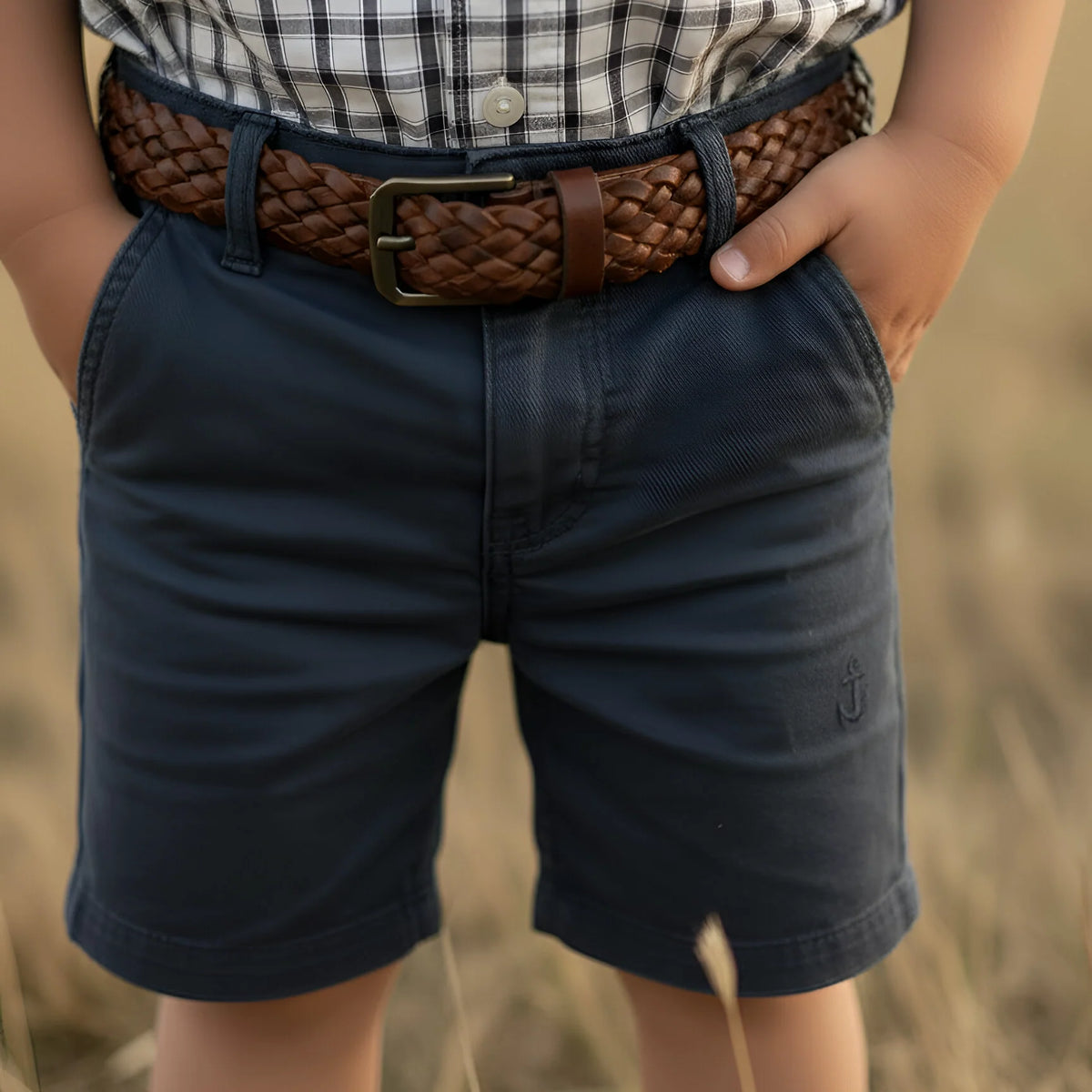 A close-up of a child wearing dark navy shorts, a brown braided belt, and a black-and-white plaid shirt, standing outdoors against a blurred tan grassy background.