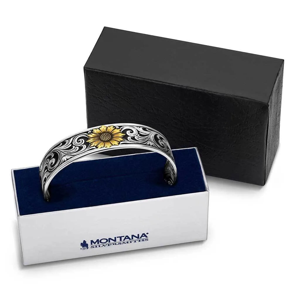 A silver-toned cuff bracelet with an engraved floral pattern and a yellow sunflower motif, displayed in an open white Montana Silver Mines gift box with a dark blue interior, with a black presentation box behind it on a white background.