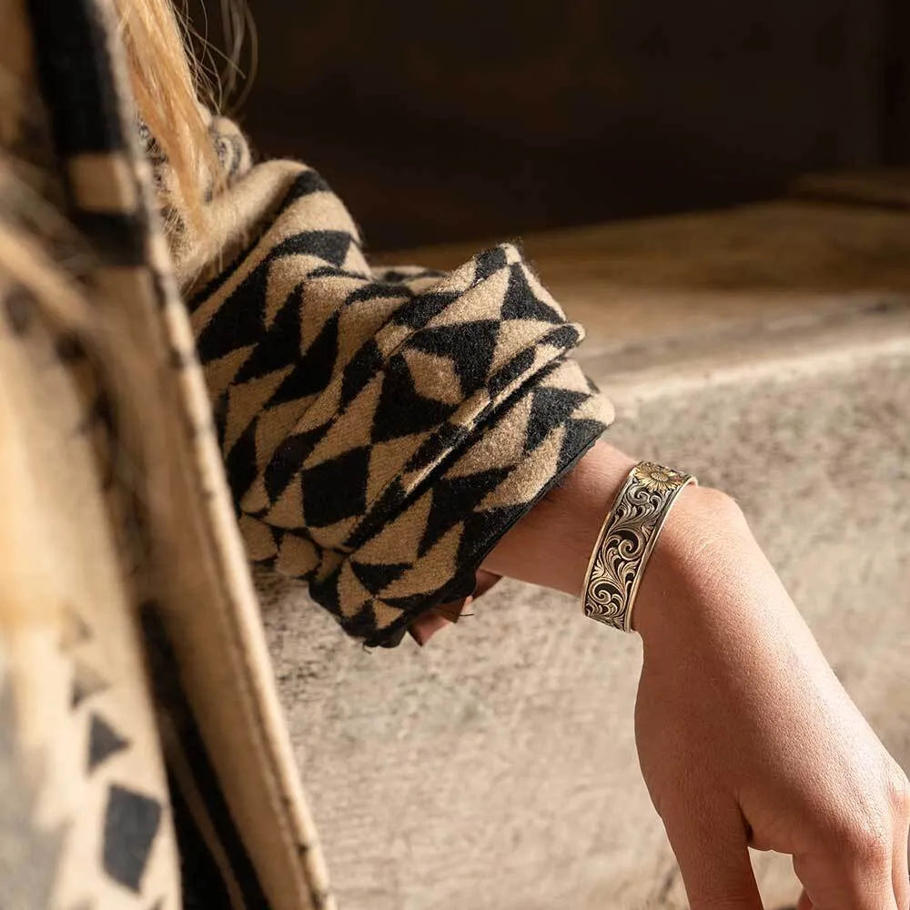A close-up of a person's wrist wearing a wide, ornate silver-toned cuff bracelet with intricate scrollwork, paired with a beige and black patterned knit sweater.