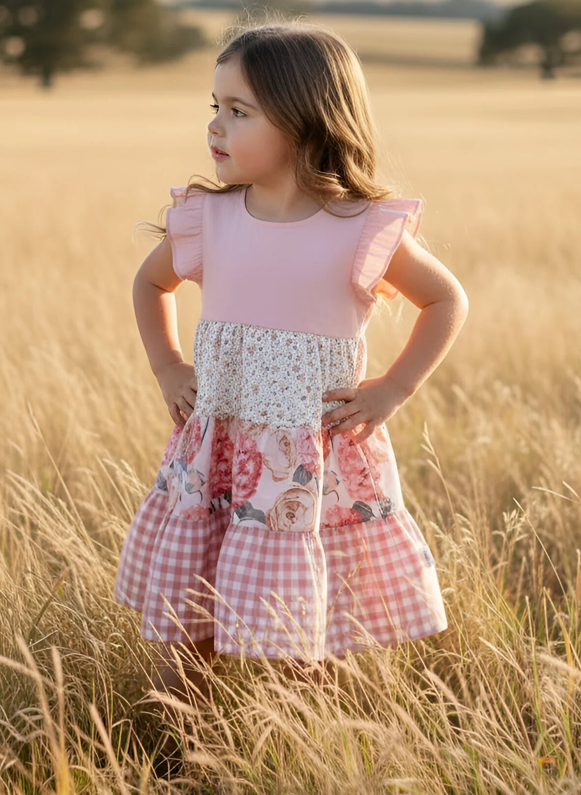 A young girl stands in a golden wheat field wearing a pink and white tiered dress with ruffle sleeves, a floral lace waist, and a pink gingham skirt.