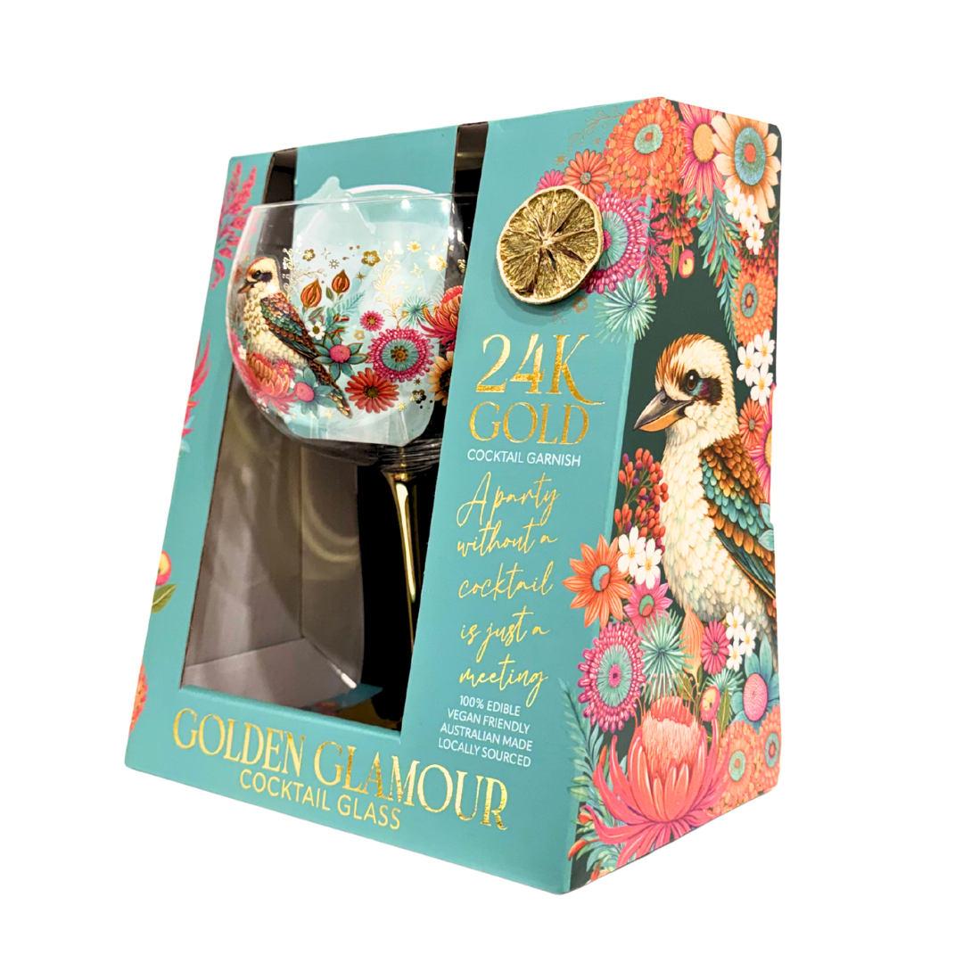 A boxed Golden Glamour 24K Gold cocktail glass set, featuring a turquoise gift box with floral artwork, a kākā pīri, and a clear window showing the decorative gold cocktail glass inside.
