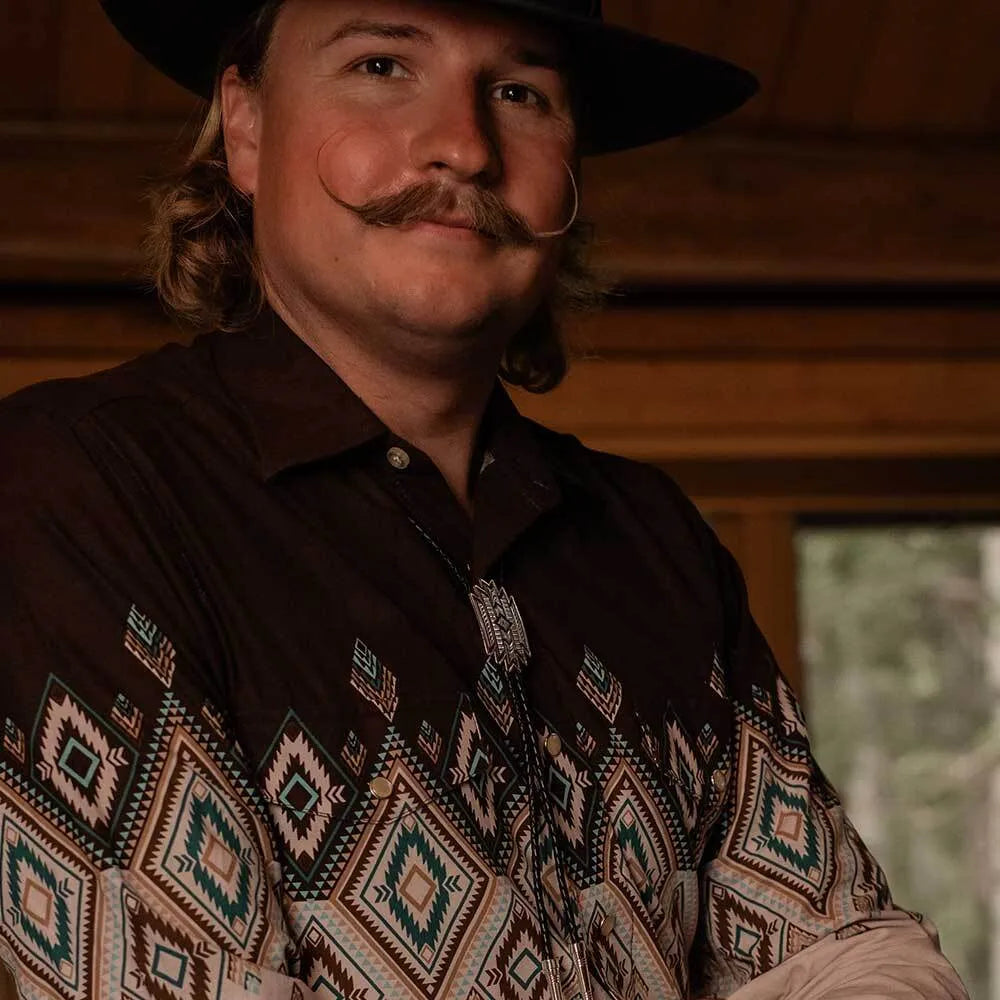 A man wearing a black cowboy hat and a dark brown long-sleeve button-up shirt with a colorful geometric southwestern pattern, posed against a rustic wooden background.