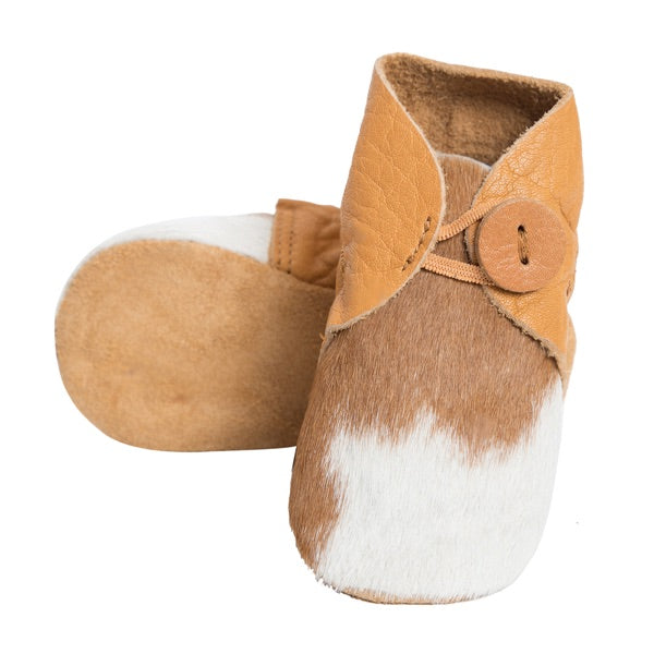 A pair of baby moccasins featuring a tan leather strap with a wooden button, a brown and white cowhide footbed, and a tan suede sole, shown on a plain white background.