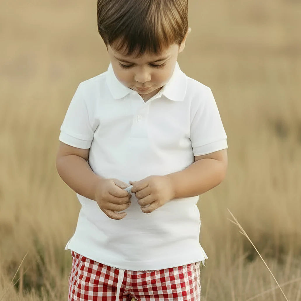 A young boy wearing a white short-sleeve polo shirt and red-and-white gingham pants standing in a dry grassy field.